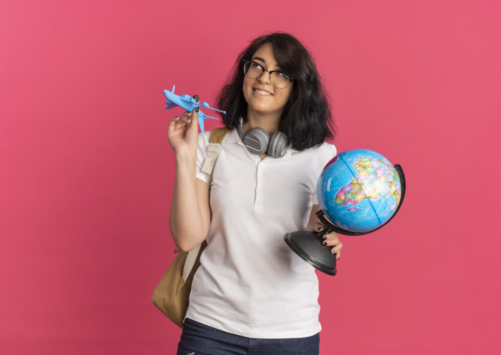 young pleased pretty caucasian schoolgirl with headphones on neck wearing glasses and back bag holds plane and globe looking up isolated on pink background with copy space