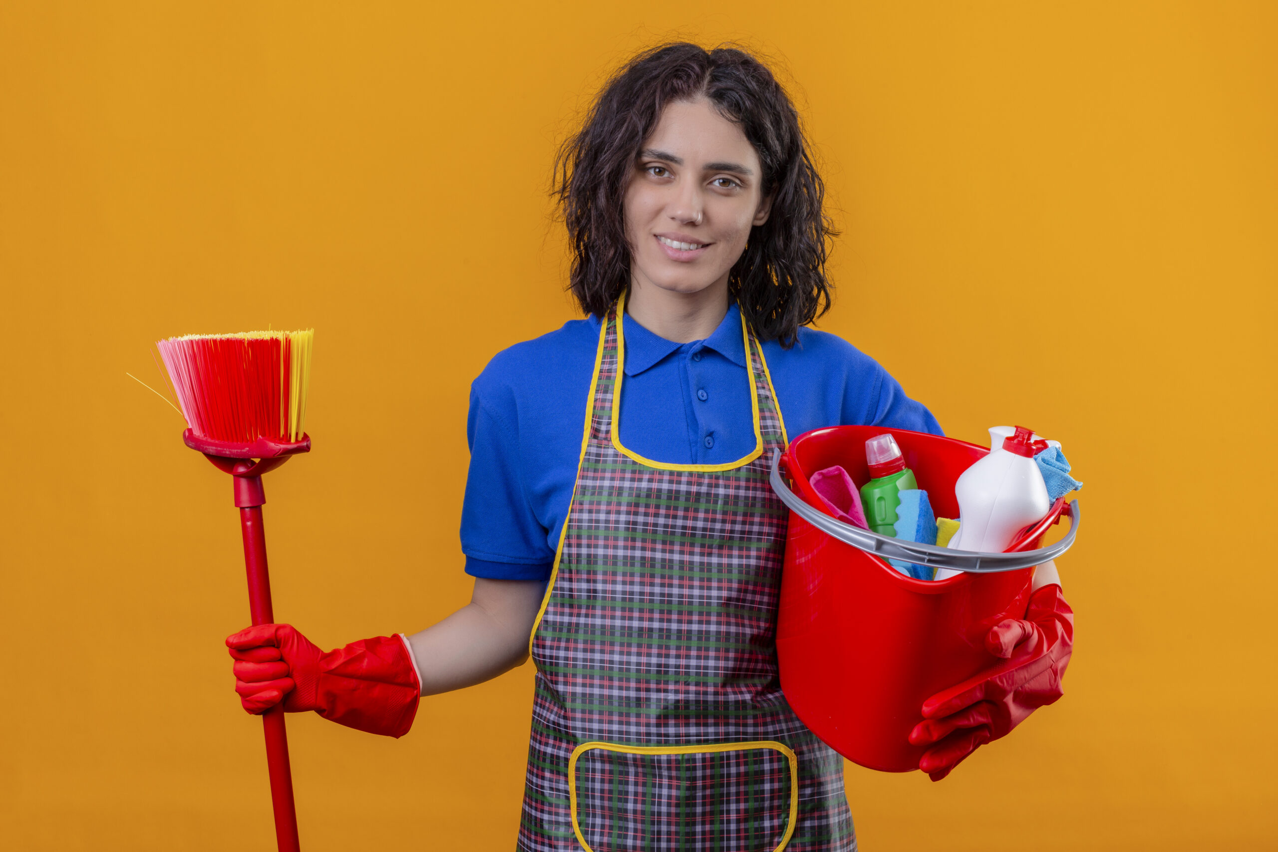 young woman wearing apron and rubber gloves holding bucket with cleaning tools and mop smiling friendly standing over orange background
